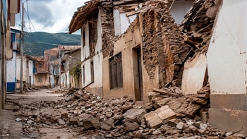 Una calle estrecha con escombros de tierra y adobes acumulados sobre el suelo frente a varias casas de estructura dañada