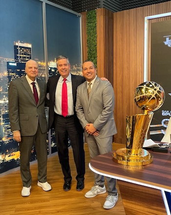 Tres hombres con traje de pie en un estudio de televisión junto a una mesa con un trofeo dorado de baloncesto
