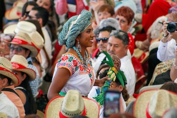 Escenario con once bailarines en trajes típicos, una bailarina de blanco al centro. Detrás, una orquesta. Al fondo, árboles y montañas. Piñas en el borde del escenario