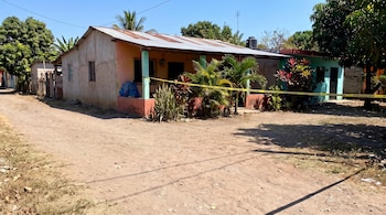 Casa de una planta con techo de chapa, fachada de colores naranja y verde, cinta de precaución amarilla, plantas, camino de tierra, árboles y cielo azul.