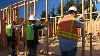 Três trabalhadores usando capacetes e roupas de proteção montam a estrutura de madeira de uma casa em construção, com céu azul e árvores ao fundo.