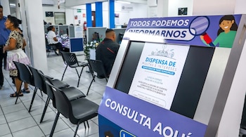 Kiosco digital interactivo con un anuncio en pantalla junto a una hilera de sillas negras en la sala de atención de una oficina municipal.