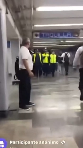 Pasillo de estación de metro. Un hombre de espaldas, camiseta blanca y pantalón negro. Grupo de personas con chalecos amarillos. Cartel azul "TAQUILLA"
