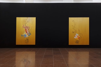 Dos cuadros con figuras invertidas y manchas de color sobre fondos dorados cuelgan en una pared negra lisa, con un suelo de baldosas marrones reflectantes