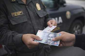 Manos intercambian un billete de cien soles peruanos y un documento; oficial de la Policía Nacional del Perú y vehículo policial de fondo.
