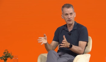 Hombre de mediana edad con camisa polo oscura y pantalón gris, sentado en silla clara, con micrófono de solapa, gesticulando. Fondo naranja