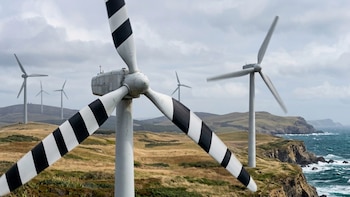 Molinos de viento con aspas blancas y franjas diagonales negras en un paisaje costero con colinas cubiertas de hierba bajo un cielo nublado