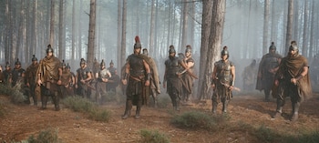 Grupo de guerreros con cascos, armaduras y capas caminando en un bosque con árboles altos y niebla