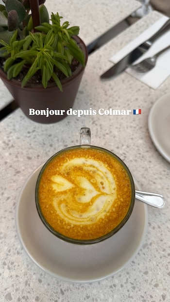 Una taza de café con un diseño en la espuma sobre una mesa junto a una pequeña planta en maceta y el texto Bonjour desde Colmar