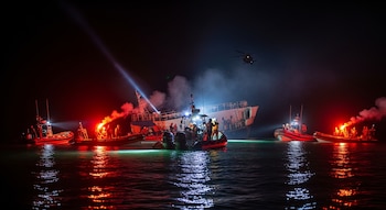 Un ferry parcialmente hundido en la noche con varias lanchas de rescate alrededor, personal con chalecos, focos de luz, bengalas rojas, humo y un helicóptero en el aire.