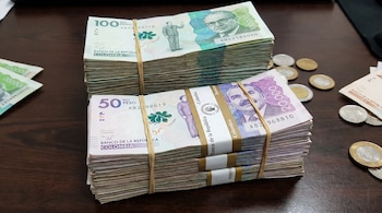 Dos grandes fajos de billetes de pesos colombianos, uno de cien mil y otro de cincuenta mil, atados con ligas, sobre una mesa oscura junto a varias monedas.