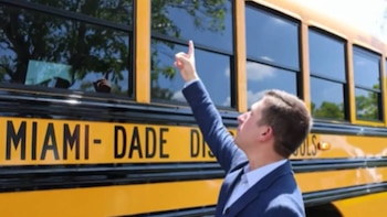 Un hombre con saco azul y camisa celeste apunta hacia el techo de un autobús escolar amarillo del distrito Miami-Dade, bajo un cielo claro