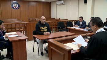 Hombre con camiseta negra y manos esposadas sentado en una mesa de madera frente a un juez y otros individuos en una sala de audiencia.