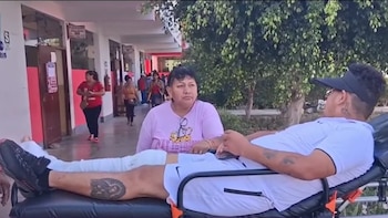 Un hombre con gorra y la pierna derecha vendada está recostado en una camilla al aire libre, con una mujer sentada a su lado; un edificio y árboles se ven al fondo