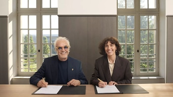 Hombre con cabello blanco y gafas de sol y mujer con cabello rizado sentados en mesa de madera, sonriendo y firmando documentos. Ventanas grandes al fondo con vista a árboles