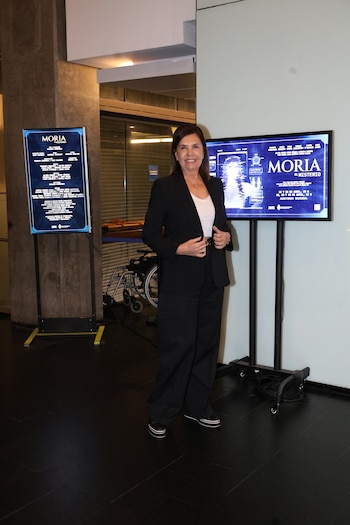 Liliana Parodi, mujer de cabello castaño y blazer negro, sonríe de pie frente a pantallas azules que promocionan "Moria, El Misterio"