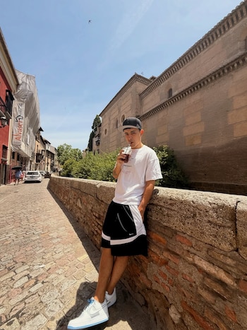 Un hombre joven con gorra, camiseta blanca y pantalones cortos negros se inclina sobre un muro de ladrillo en una calle empedrada, bebiendo de un vaso