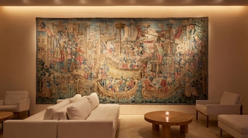 Un gran tapiz mural de escena naval o histórica domina una sala con sofás modulares blancos, sillones y una mesa de centro de madera iluminada.