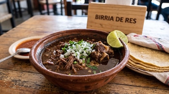 Cazuela de barro con birria de res, cilantro, cebolla y limón, junto a tortillas, salsa y un letrero de madera con la frase BIRRIA DE RES.