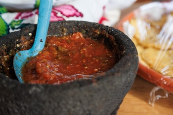 Molcajete negro de piedra lleno de salsa roja con una cuchara azul, junto a un recipiente cubierto con plástico sobre una mesa de madera