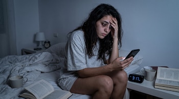 Una mujer joven con cabello oscuro sentada en una cama. Sostiene un teléfono móvil iluminado. Al lado un despertador digital marca 3:17 AM. Hay libros y tazas.