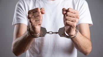 Un hombre viste una camiseta blanca y sostiene sus manos esposadas con los puños cerrados sobre un fondo gris.