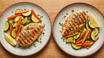 Vista superior de dos platos sobre madera con pechugas de pollo marcadas a la plancha, rodajas de zucchini, bastones de zanahoria y limón.