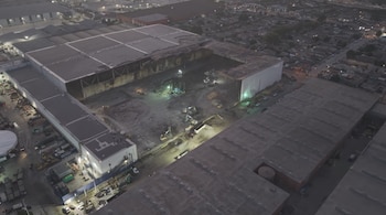 Vista aérea de un gran edificio industrial con una sección interior quemada, excavadoras, maquinaria, luces, otros edificios, calles y vehículos