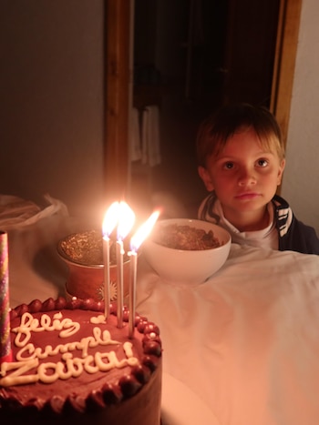 Torta de chocolate con inscripción "Feliz cumple Zaira" y cuatro velas encendidas. Un niño, un mate y un cuenco con cereales