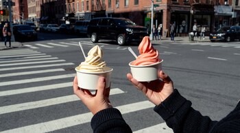 Dos manos sostienen dos vasos de helado de crema suave, uno amarillo y otro rosado, con cucharas blancas, en un fondo de calle con cruce peatonal y edificios.
