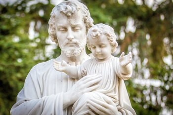 Estatua de mármol blanco de San José con barba y el Niño Jesús. Ambos tienen vestimenta religiosa y están sobre un fondo verde borroso