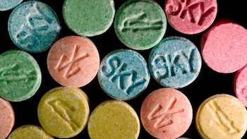 Un surtido de píldoras redondas de colores (azul, verde, rosa, amarillo) con marcas como "SKY", un cocodrilo y otros símbolos, sobre un fondo negro