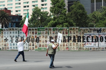 Dos hombres caminan por una calle. Detrás, un muro cubierto con fotos de personas y el texto "+130,000 DESAPARECIDOS". Uno sostiene una bandera mexicana, el otro un cartel blanco