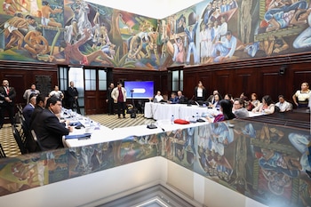 Personas sentadas alrededor de mesas con manteles blancos en un salón con micrófonos, pantallas y murales en las paredes