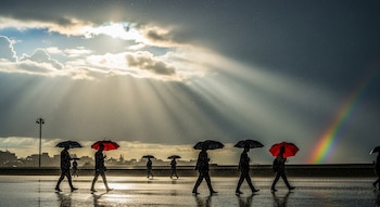 Siluetas de personas caminan con paraguas bajo la lluvia y sol. Rayos solares y un arcoíris visible. Horizonte urbano en el fondo.