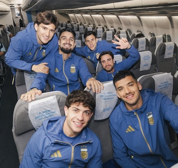 Seis jóvenes de la selección argentina posan sonrientes en el interior de un avión, vistiendo chándales azules con el logo de la AFA