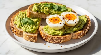 Un plato blanco contiene tostadas integrales con aguacate untado y dos mitades de huevo cocido, sobre una mesa de mármol blanco bajo luz natural.