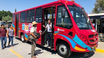 Un hombre con sombrero de paja sostiene un guajolote negro frente a un autobús rojo Binnibus, mientras habla con el conductor en la entrada.
