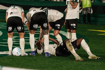Jugador de fútbol tendido en el césped, rodeado por tres compañeros de equipo. Un balón de fútbol blanco y azul está en el suelo
