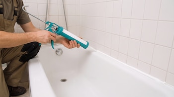 Manos de un trabajador utilizando una pistola de calafateo verde para aplicar sellador blanco entre una bañera y una pared de azulejos.