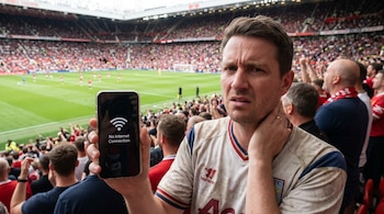 Primer plano de un hombre frustrado con camiseta de fútbol, mostrando un celular con mensaje 'No Internet Connection' en un estadio lleno de gente.