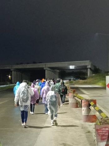 Personas de espalda caminando de noche por una carretera junto a un puente, con impermeables de colores y mochilas