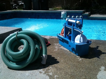 Una manguera verde enrollada y una caja de herramientas azules con cepillos y botellas de limpieza, ubicadas al borde de una piscina con agua azul