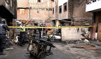 Calle con escombros y restos quemados, un carro de mano calcinado, oficial de policía, hombre con chaleco amarillo, grupo de personas y cinta de seguridad amarilla