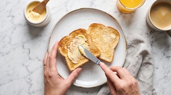 Manos que untan mantequilla con un cuchillo en una tostada dentro de un plato, junto a una taza de café y un recipiente.