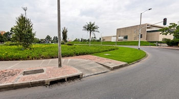 Una calle curva con acera, un área de césped con árboles, un semáforo y un edificio moderno al fondo bajo un cielo nublado.