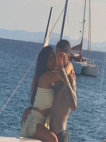 Un hombre y una mujer se abrazan en un barco. El hombre sujeta un teléfono naranja. Al fondo se ven el mar, montañas y varios veleros