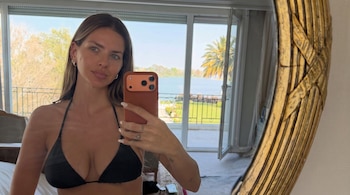 Una mujer con traje de baño negro sostiene un teléfono para tomarse una foto frente a un espejo redondo de marco dorado con un ventanal detrás.