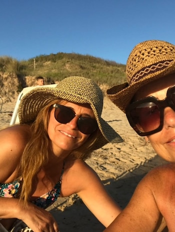 Dos mujeres con sombreros de paja y gafas de sol se encuentran en una playa de arena; al fondo, dunas y un cielo azul claro
