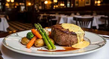 Un medallón de Châteaubriand cubierto con salsa bearnesa, acompañado de espárragos, zanahorias y patatas, servido en un plato blanco con fondo borroso de un restaurante.